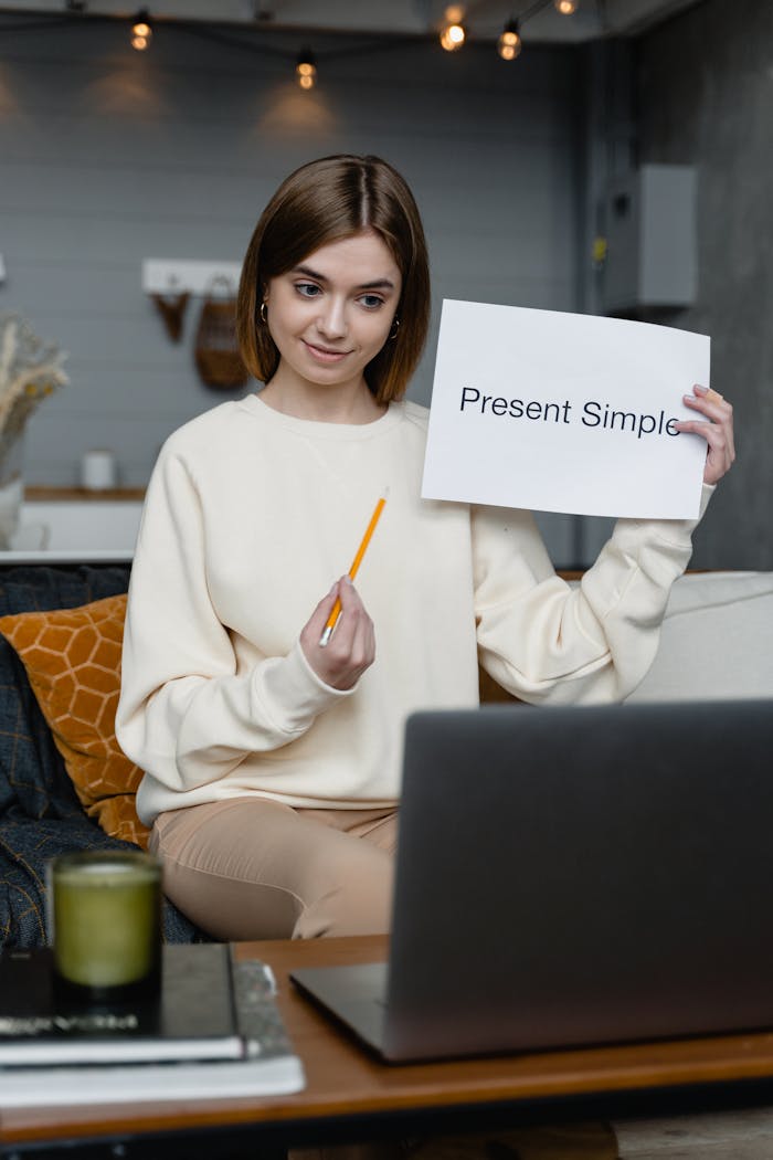 Exam Format: What Does Each Test Actually Look Like? Woman teaching English online holding 'Present Simple' flashcard, engaging in video call.